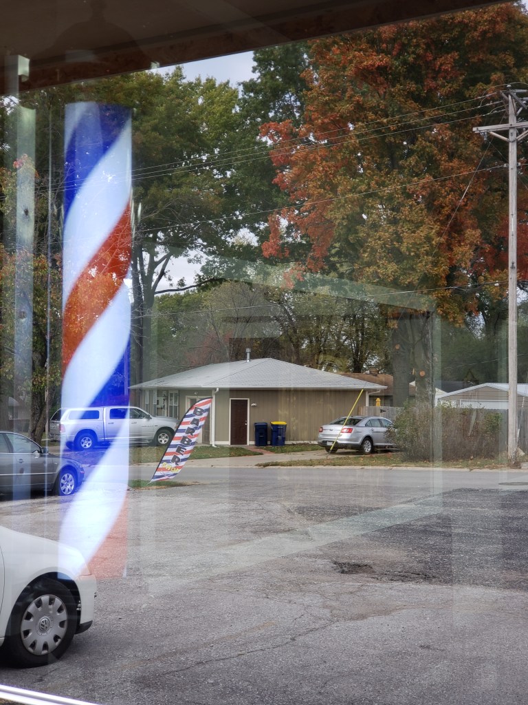 The reflection of the Barbershop banner in the front window of the shop. The lit barber pole inside is visible.