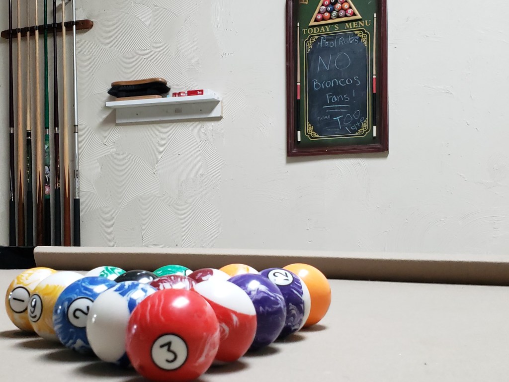 The pool setup in the back of the shop. The balls are on the table, cues are in the rack on the wall, and a chalkboard says "Pool rules: No Broncos fans! Too late."