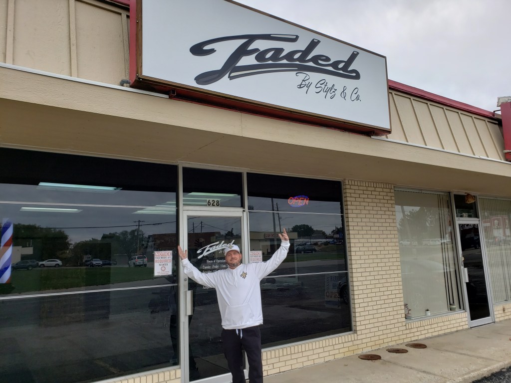 Stylz smiling with his arms raised in celebration in front of the shop.