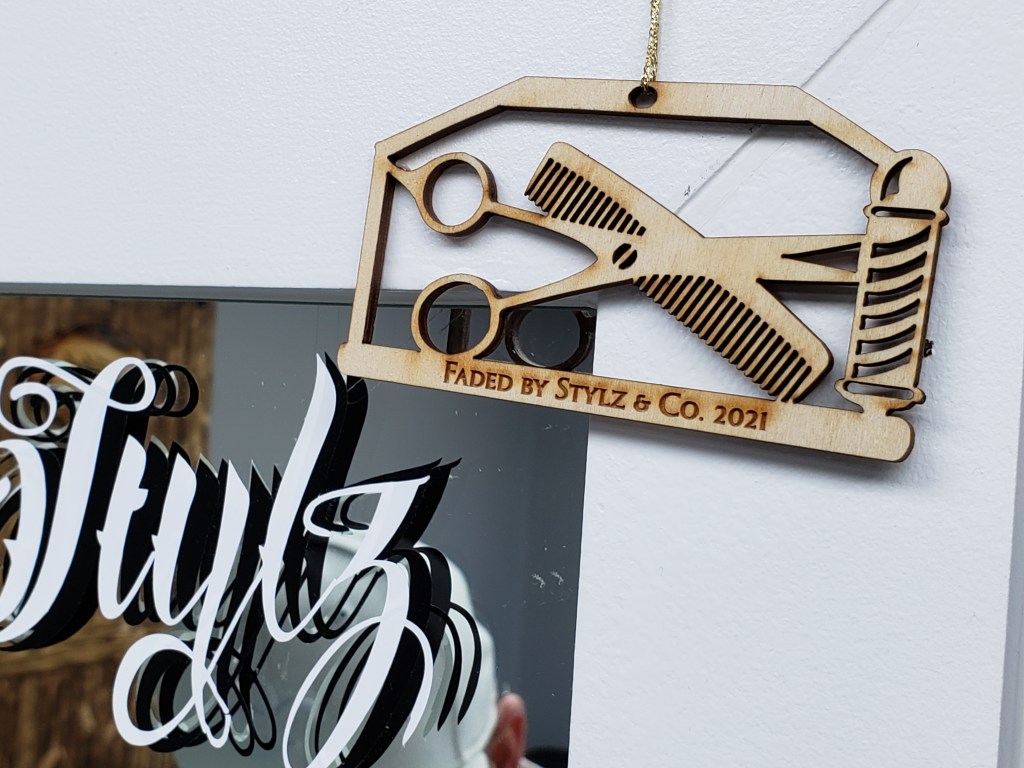 A wooden die-cut ornament with a barber pole and crossed scissors and comb and the words Faded By Stylz & Co. 2021.