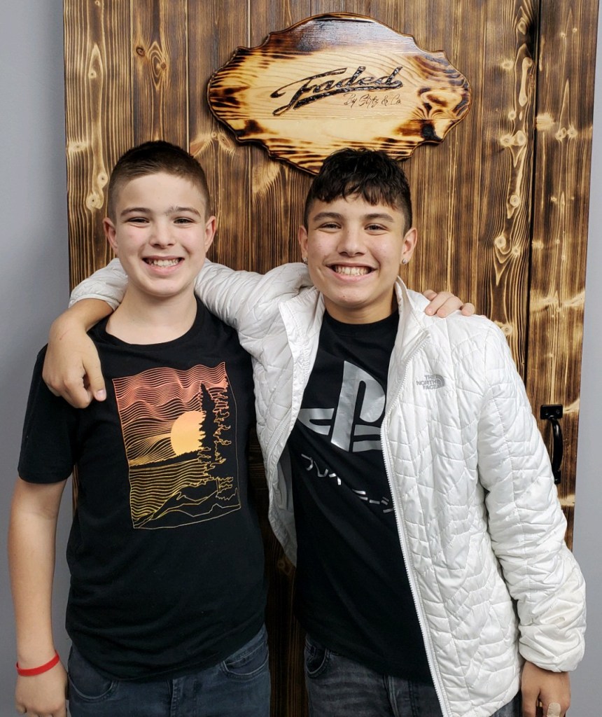 Two adolescent boys with freshly cut hair standing by the Faded wooden sign.